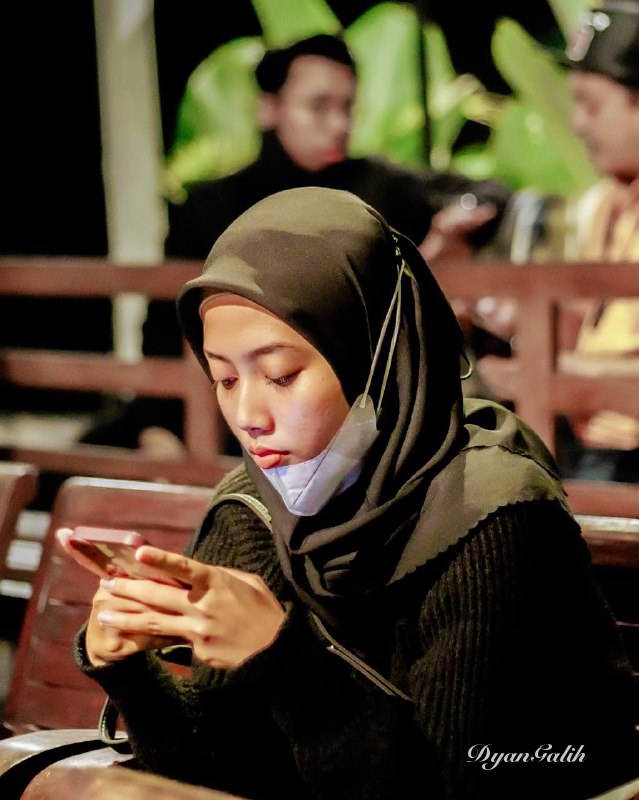 A young woman wearing a hijab is playing with her gadget against blur background. Gear: Canon EOS Rp. Lens: Canon 50mm. Model: -. Spot: Warung Kulinan.